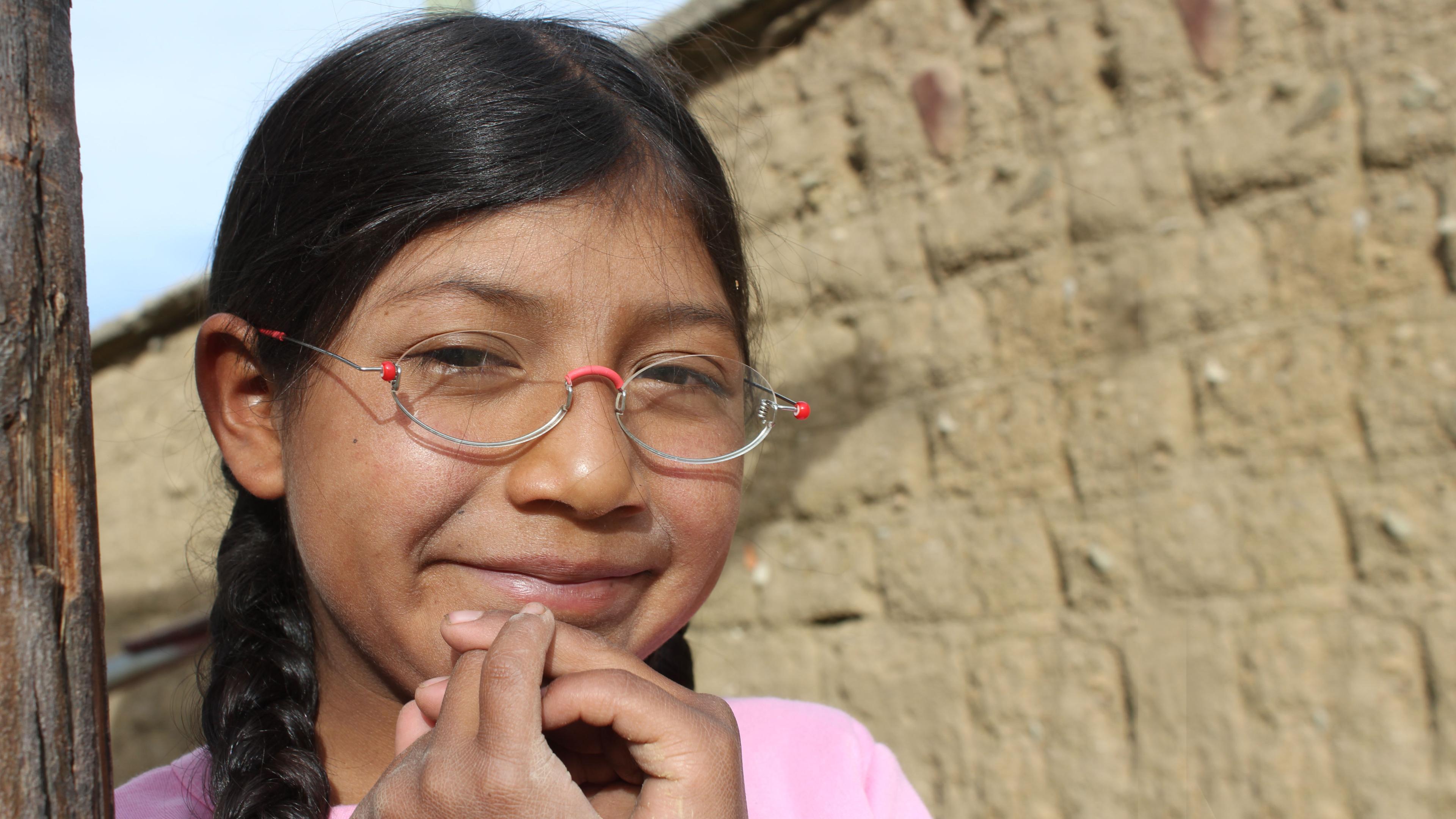 Mädchen aus Bolivien mit EinDollarBrille, grinst und hält sich die gefalteten Hände ans Kinn